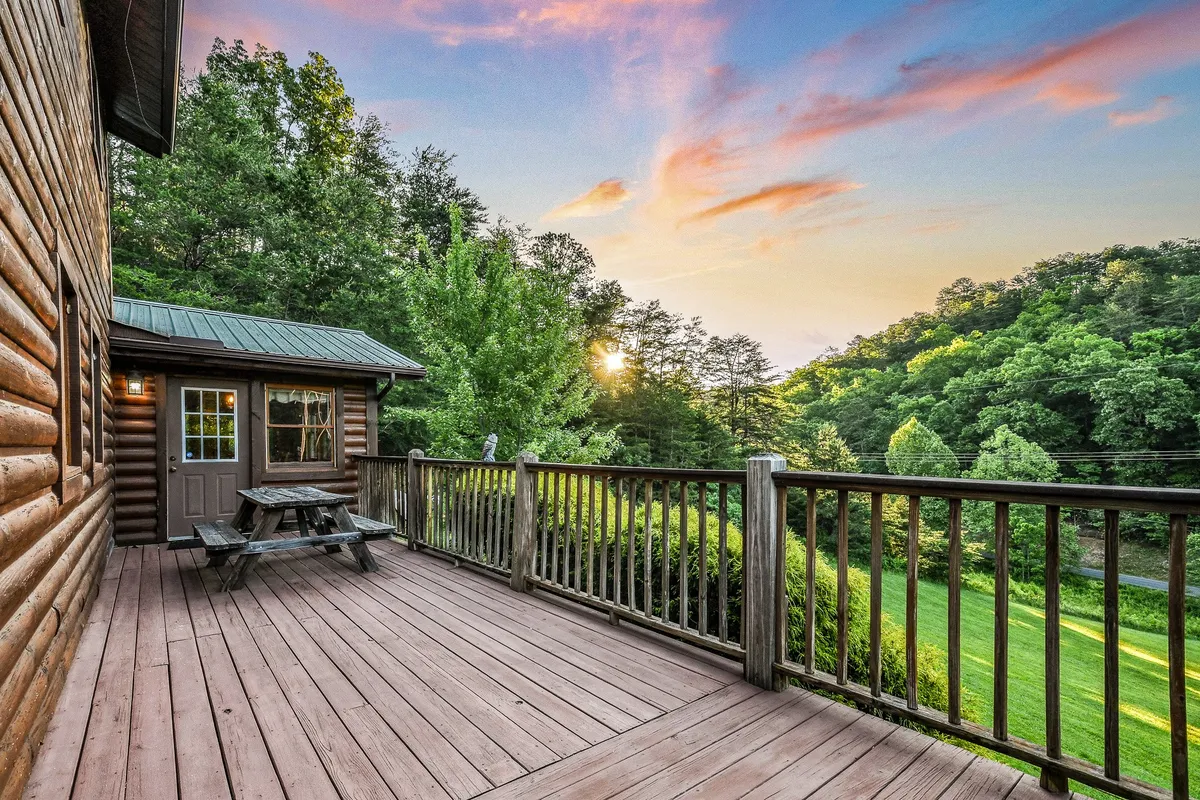 A wide deck with outdoor furniture overlooks surrounding trees and a vibrant sunset sky. The setup is perfect for outdoor meals, barbecues, or simply enjoying the peaceful atmosphere with family and friends.