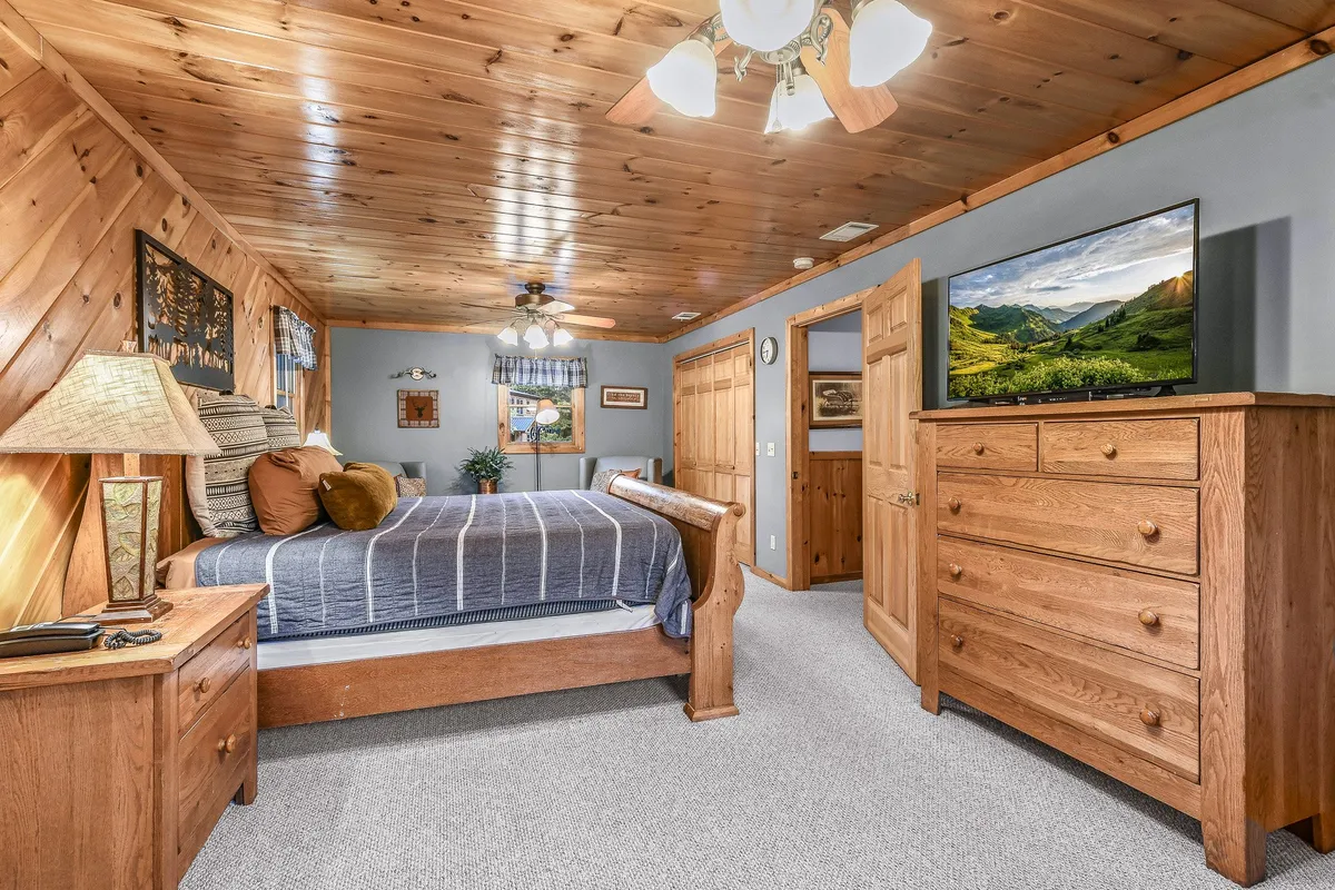 A serene bedroom scene with a bed dressed in striped bedding, a wooden dresser, a television displaying a tranquil landscape, and soft lamp lighting. The room is bright with natural sunlight filtering in, creating a peaceful and comfortable ambiance.