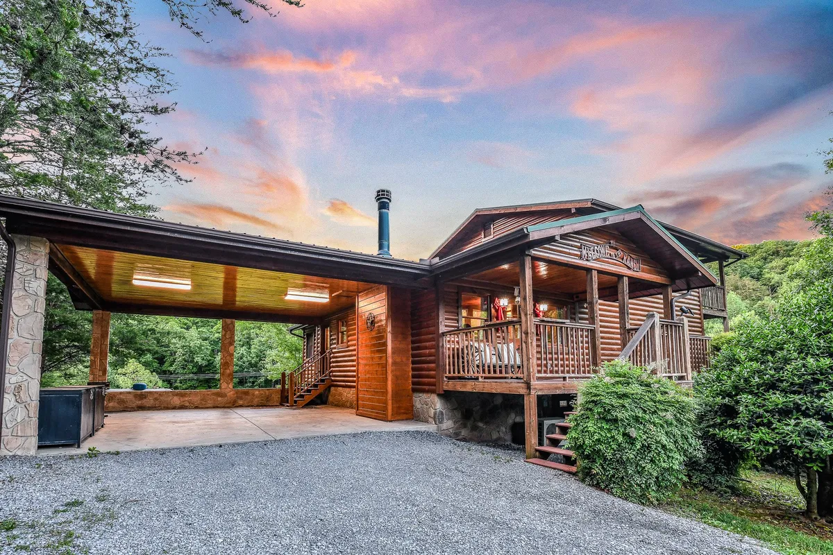 A wide exterior shot of The Ranch's log cabin structure at sunset, surrounded by lush greenery. The carport and porch are visible, with the enchanting colors of the sky suggesting the close proximity to nature and the sense of arrival at a mountain retreat.