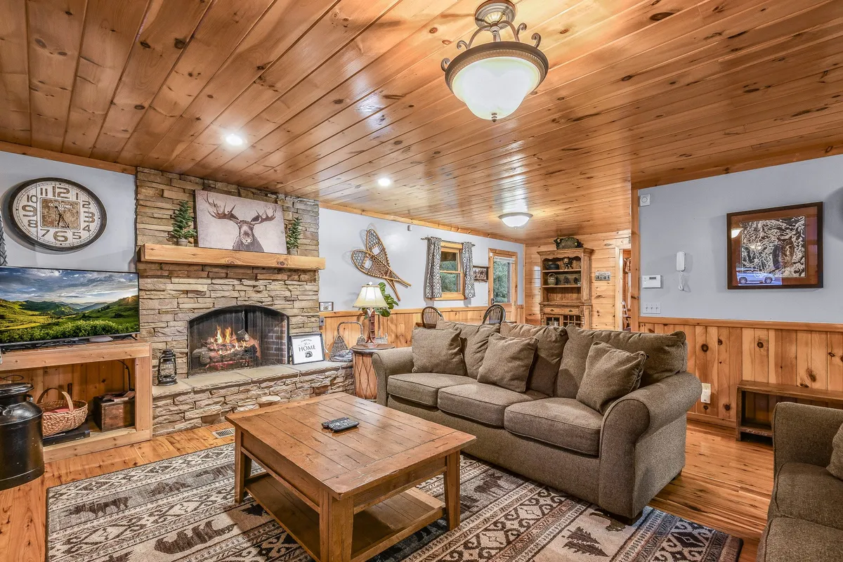 A warm, open living area with a fireplace, a cozy sofa, a wooden coffee table, a wooden ceiling, and decorative wall art. The arrangement invites group gatherings and comfortable relaxation against a backdrop of rustic charm.