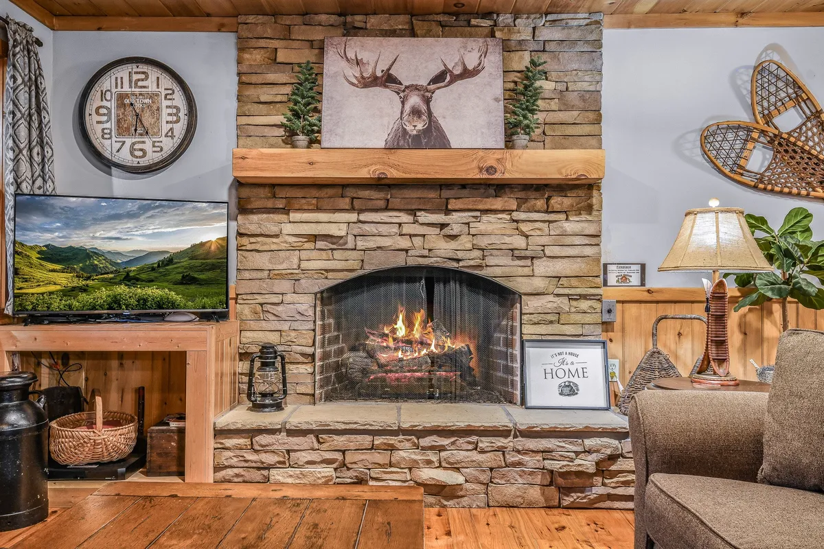 A cozy living room featuring a stone fireplace, a television displaying a scenic landscape, a comfortable sofa, a wooden coffee table, and inviting ambient lighting from a table lamp. The room exudes warmth with rustic accents and moose-themed artwork above the mantelpiece.