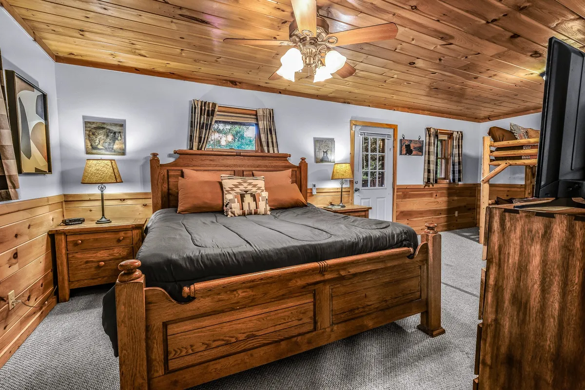 A warmly appointed bedroom featuring a large wooden king bed alongside a queen-over-queen bunk bed. The room is well-lit, decorated with soft lighting from decorative lamps, and includes framed artwork. The atmosphere feels inviting and perfect for families or groups, with ample space and comfortable bedding.