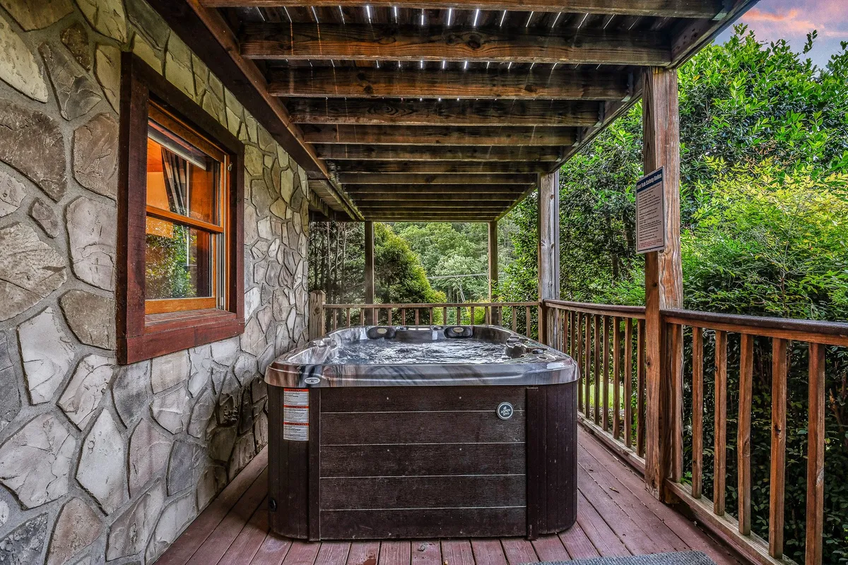 This image showcases a tranquil outdoor hot tub on a wooden deck, surrounded by lush greenery and a stone wall. The scene suggests a cozy, peaceful environment ideal for soaking and socializing while enjoying nature's views.