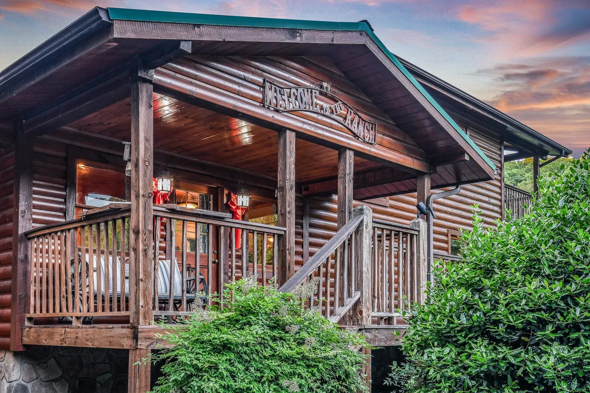 This image shows the inviting exterior of a log cabin, featuring a welcoming porch with a railing and a visible welcome sign. The setting radiates an atmosphere of relaxation and togetherness, and the porch offers space for groups to gather and enjoy the outdoors.