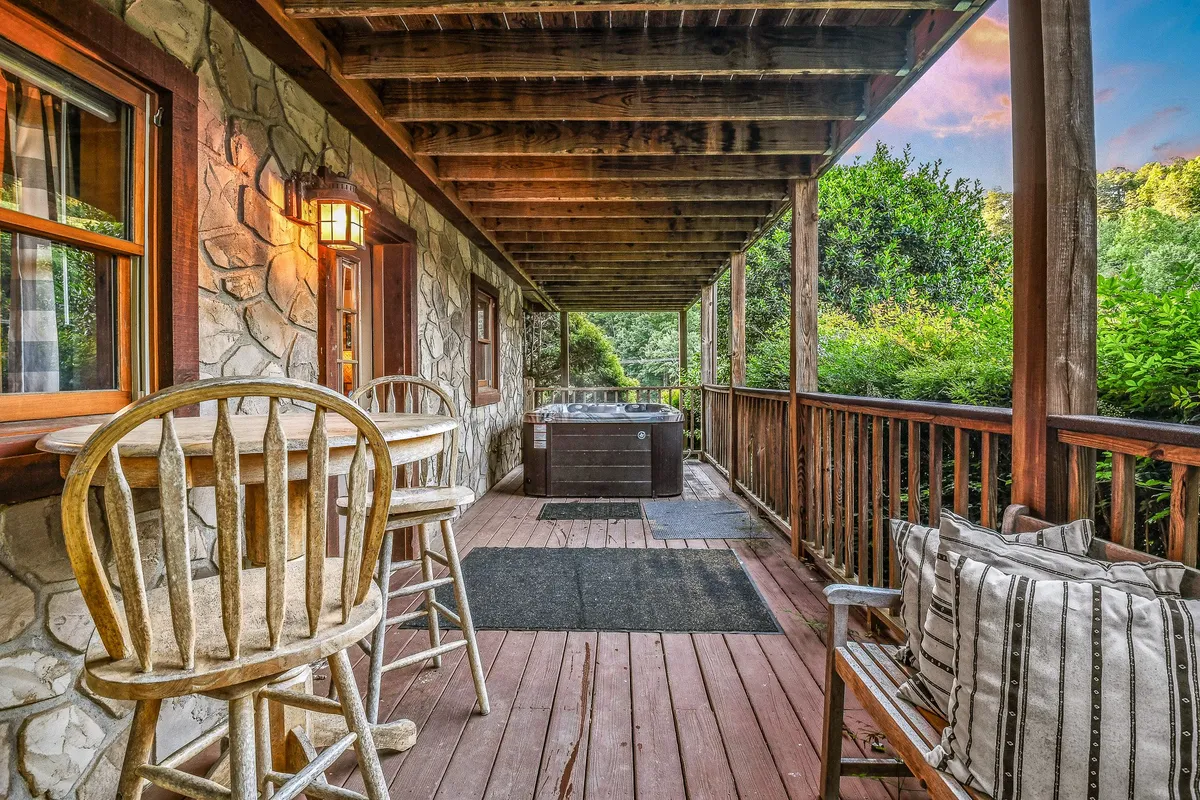 A serene exterior scene featuring a hot tub on a wooden deck, surrounded by a stone wall, wooden railing, and nearby wooden chairs. The area looks inviting for relaxing under the stars or gathering with friends. The backdrop hints at natural privacy and scenic views, making it ideal for unwinding after a night out.