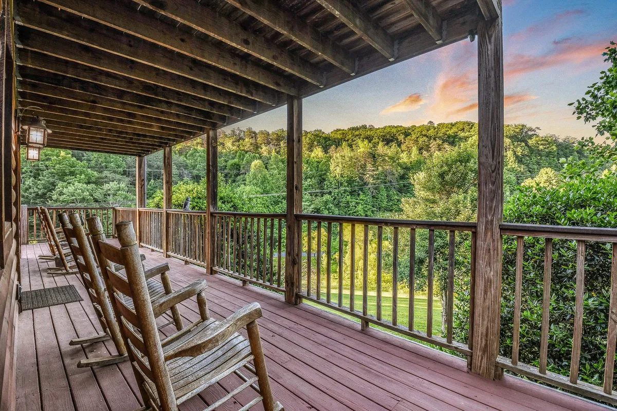 A pair of classic rocking chairs face out from a covered deck, framed by a wooden railing, lush greenery, and a glowing sunset sky. The scene exudes comfort and invites guests to relax, read, or socialize while enjoying the mountain atmosphere.