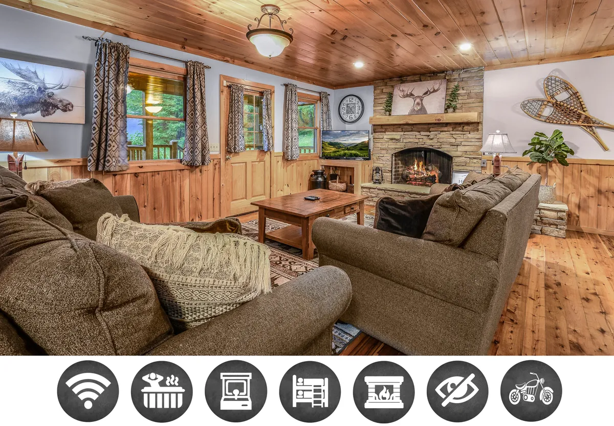 The image showcases a cozy cabin living room with a stone fireplace as the centerpiece, surrounded by a plush sofa and rustic wooden coffee table. Warm, inviting lighting and tasteful decorative wall art create a welcoming atmosphere, perfect for gathering, relaxing, or enjoying a movie night with friends and family.