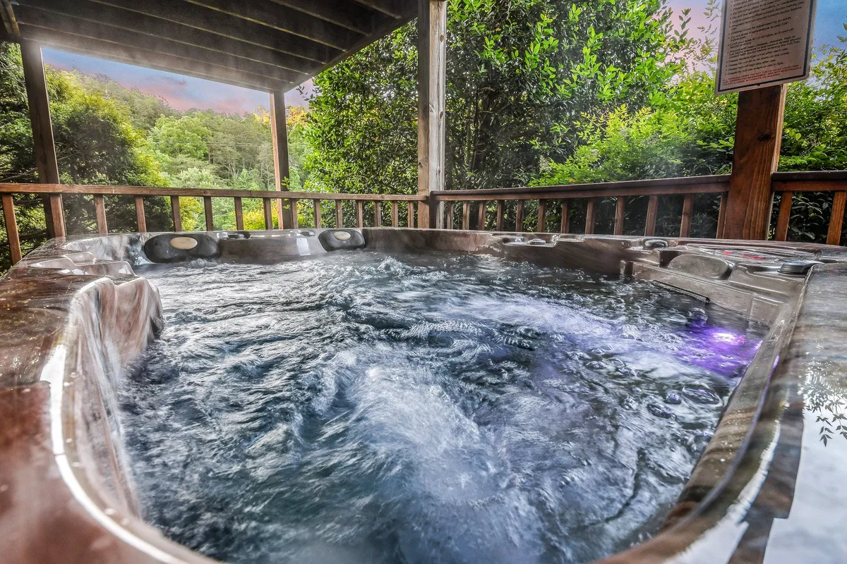 A bubbling hot tub sits on a wooden deck, steam rising against a lush, green backdrop. The scene is inviting, tranquil, and embodies relaxation while immersed in nature. The hot tub is a focal point, encouraging guests to unwind and soak after a day in the mountains.