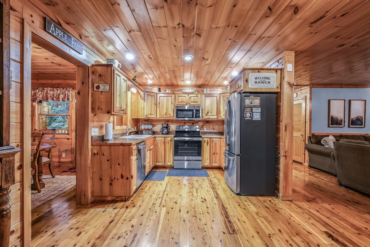 An open-plan view capturing the kitchen, wooden dining table with chairs, and adjacent living area. The kitchen area is appointed with stainless steel appliances, wooden cabinetry, and decorative touches, while the dining space provides ample seating for shared meals.