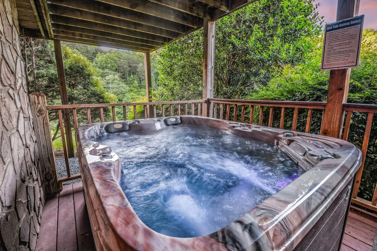 A tranquil view of an outdoor hot tub set against a backdrop of lush greenery, with a safety rules sign nearby. The open sky above hints at the opportunity for stargazing or unwinding after an eventful day.